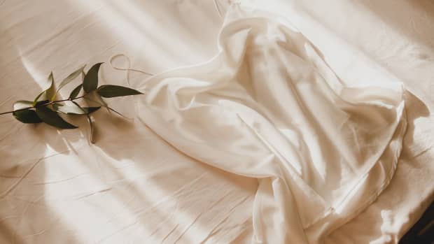 A silk white blouse with thin straps lies on a crumpled sheet with a branch of eucalyptus in the morning sun.