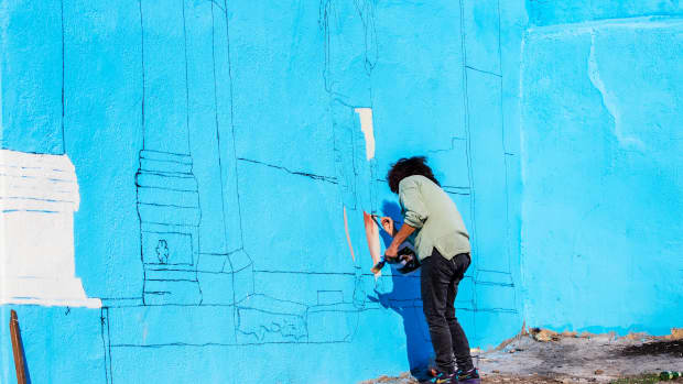 Izmir, Turkey, October 27, 2021: Black-haired woman painting a wall paints the blue building in front of the ancient city of Agora in Çankaya district. Unidentified mural painter. Remote shooting.