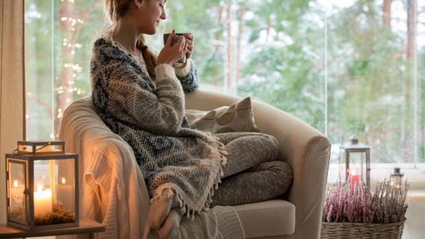 Young beautiful woman sitting home in the chair by the window with cup of hot coffee wearing knitted warm sweater. Cozy room decorated with lanterns and candles.